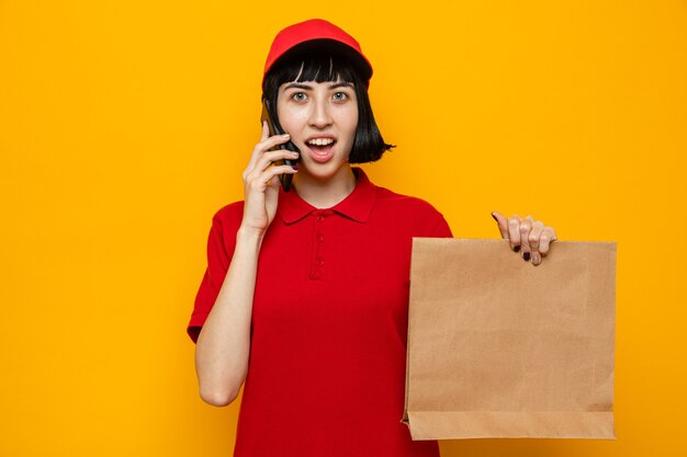 Zaskoczona młoda kaukaska kobieta dostarczająca jedzenie, trzymająca papierowe opakowanie żywności i rozmawiająca przez telefon