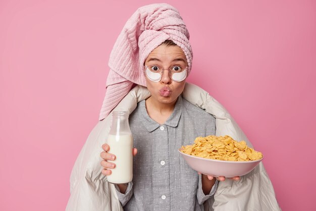Zaskoczona młoda kobieta ma zdrowe śniadanie trzyma miskę płatków zbożowych i butelkę mleka nosi piżamę i plastry kosmetyczne ręcznik kąpielowy przykryty kocem trzyma usta złożone na białym tle na różowym tle