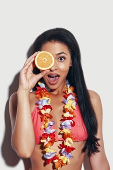 Zaskoczona młoda kobieta w bikini zakrywająca oko plasterkiem pomarańczy i uśmiechnięta stojąc na szarym tle
