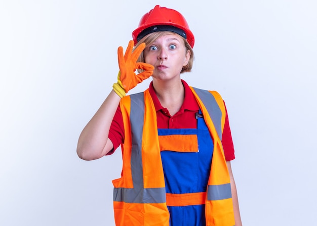 zaskoczona młoda konstruktorka w mundurze w rękawiczkach pokazująca gest ciszy na białej ścianie