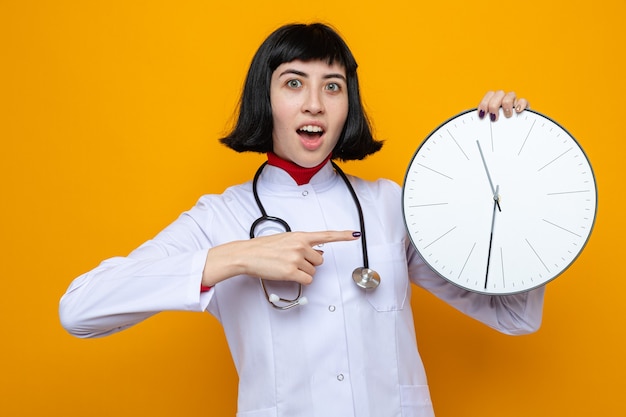 Zaskoczona młoda ładna kaukaska dziewczyna w mundurze lekarza, trzymająca stetoskop i wskazująca na zegar