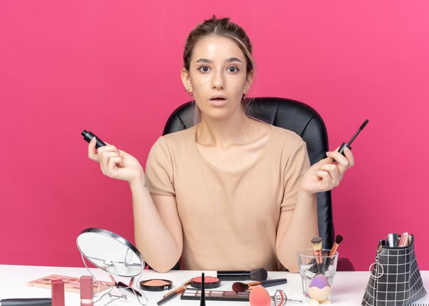 Bezpłatne zdjęcie zaskoczona młoda piękna dziewczyna siedzi przy stole z narzędziami do makijażu, trzymając pudrowy rumieniec rozprowadzający ręce na białym tle na różowym tle