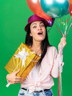 Zaskoczona młoda piękna kobieta w kapeluszu imprezowym, trzymająca balony trzymające pudełko na białym tle na zielonej ścianie