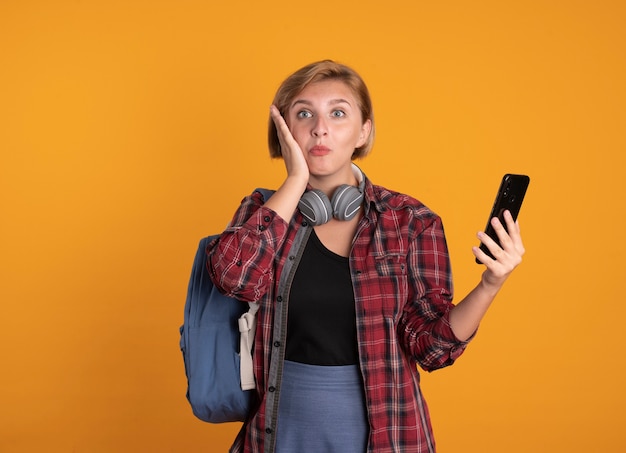 Zaskoczona młoda słowiańska studentka ze słuchawkami w plecaku kładzie rękę na twarzy i trzyma telefon
