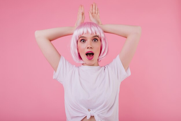 Zaskoczona modna kobieta pozuje na pastelowej ścianie. Ekstatyczna dziewczyna w różowym peruke bawi się podczas sesji zdjęciowej w domu