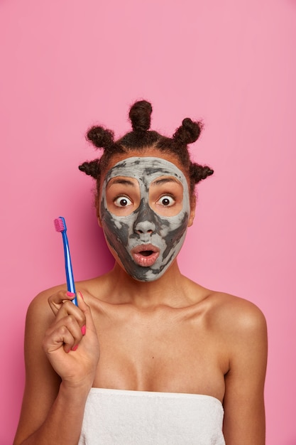Bezpłatne zdjęcie zaskoczona przestraszona kobieta wpatruje się w zatkane oczy, trzyma szczoteczkę do zębów, idzie do czyszczenia zębów po zdjęciu maski upiększającej z twarzy, dba o urodę i higienę, nosi ręcznik kąpielowy na nagim ciele