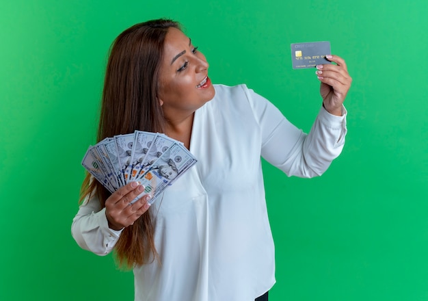 Bezpłatne zdjęcie zaskoczona przypadkowa kaukaska kobieta w średnim wieku trzymająca gotówkę i patrząca na kartę kredytową w dłoni na zielonym tle