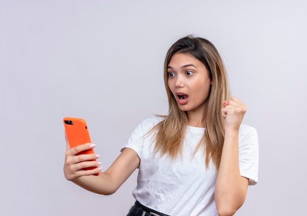 Zaskoczona śliczna młoda kobieta w białej koszulce patrząc na telefon komórkowy podnosząc zaciśniętą pięść na białej ścianie