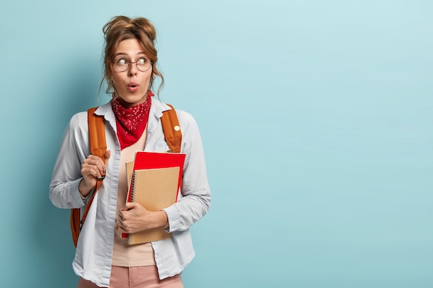 Zaskoczona uczennica z plecakiem, trzymająca spiralny notatnik, czerwony podręcznik, zszokowana sesją w przyszłym tygodniu, nosi okrągłe okulary