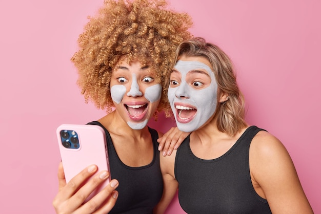 Zaskoczone Szczęśliwe Młode Kobiety Patrzą Na Ekran Smartfona Sprawiają, że Selfie Trzymają Otwarte Usta, Nakładają Błotną Maskę Na Twarz Obok Siebie, Ubrane W Zwykłe Czarne Koszulki Na Białym Tle Na Różowym Tle