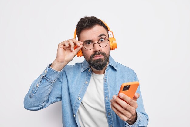 Zaskoczony brodaty mężczyzna patrzy uważnie, nosi okrągłe okulary, używa bezprzewodowych słuchawek do słuchania muzyki, trzyma, pobiera ze smartfona, piosenkę z listy odtwarzania, ma na sobie zwykłą koszulę