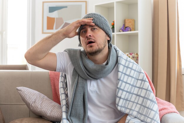 Zaskoczony chory słowiański mężczyzna z szalikiem na szyi w czapce zimowej owiniętej w kratę, kładący dłoń na czole, siedzący na kanapie w salonie
