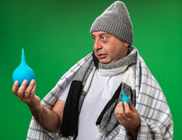 Bezpłatne zdjęcie zaskoczony dorosły chory kaukaski mężczyzna z szalikiem na szyi w czapce zimowej owiniętej w kratę trzymający i patrzący na lewatywy odizolowane na zielonej ścianie z kopią miejsca