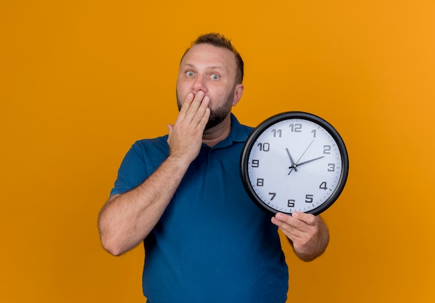 Bezpłatne zdjęcie zaskoczony dorosły człowiek słowiański gospodarstwa zegar kładąc rękę na ustach patrząc