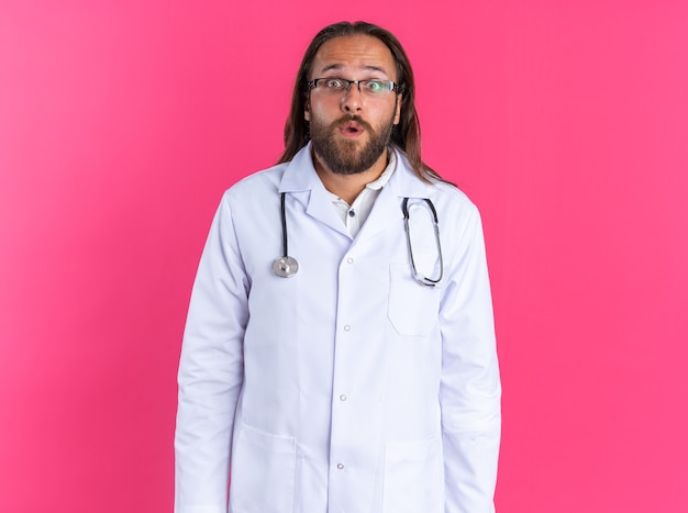 Zaskoczony dorosły lekarz mężczyzna ubrany w szatę medyczną i stetoskop w okularach, patrzący na kamerę odizolowaną na różowej ścianie