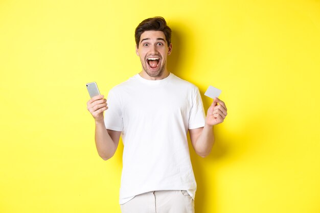 Zaskoczony facet trzyma smartfon i kartę kredytową, zakupy online w czarny piątek, stojąc na żółtym tle.