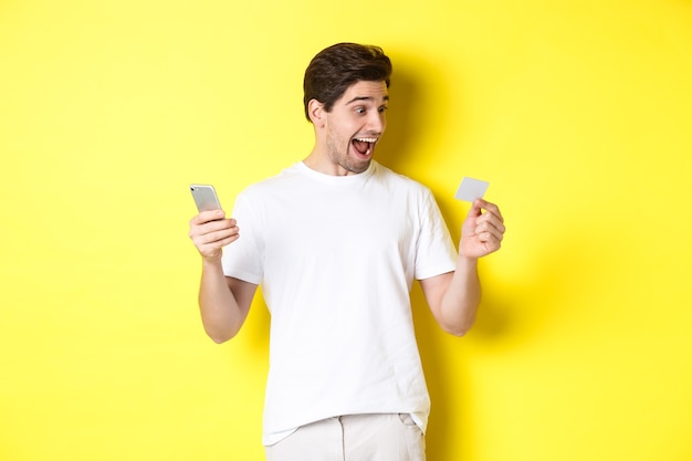 Zaskoczony facet trzyma smartfon i kartę kredytową, zakupy online w czarny piątek, stojąc na żółtym tle.
