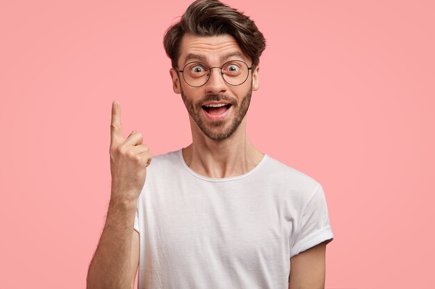 Zaskoczony hipster z modną fryzurą, ma intrygujące zszokowane spojrzenie, wskazuje palcem wskazującym w górę, nosi zwykłą białą koszulkę i okulary, odizolowane na różowej ścianie