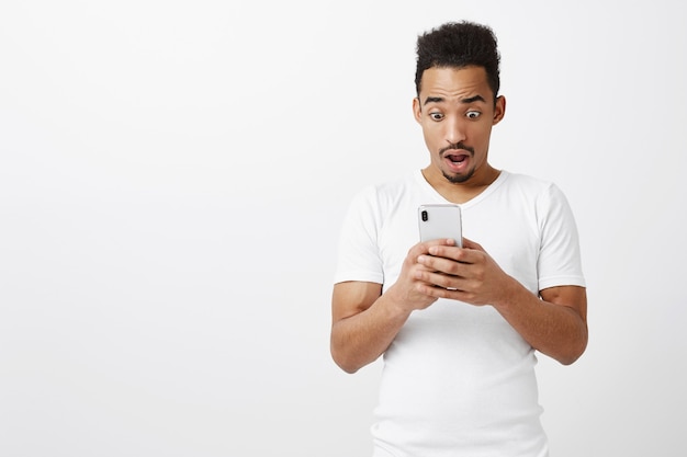 Zaskoczony i zdumiony Afroamerykanin sapiąc, patrząc na ekran smartfona zdziwiony wielką wiadomością, niesamowitą aplikacją