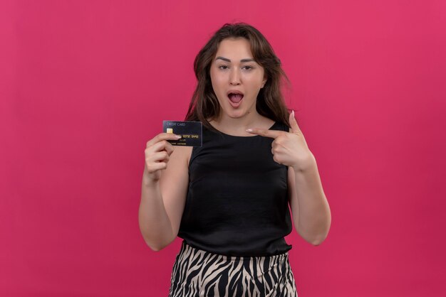 Zaskoczony kaukaski dziewczyna ubrana w czarny podkoszulek trzyma kartę bankową i wskazuje kartę bankową na różowym tle