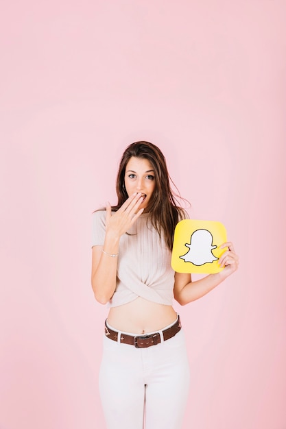 Bezpłatne zdjęcie zaskoczony kobieta trzyma ikonę snapchat na różowym tle