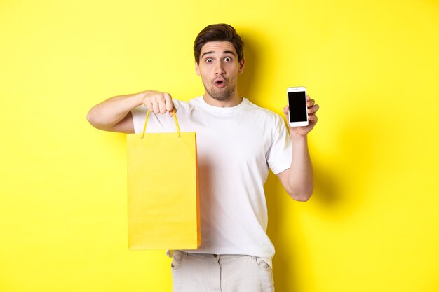 Zaskoczony mężczyzna trzyma torbę na zakupy i pokazuje ekran smartfona, koncepcja bankowości mobilnej i osiągnięć aplikacji, żółte tło.
