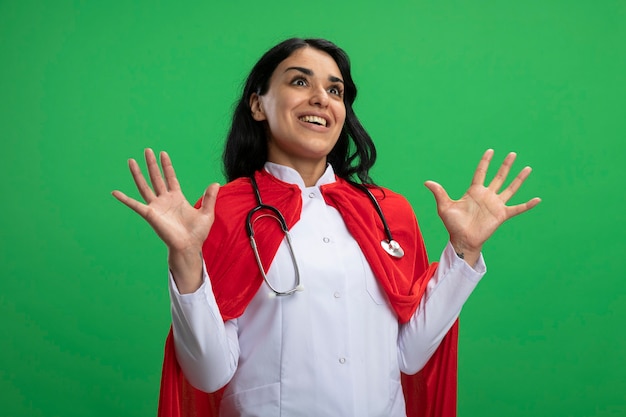 Bezpłatne zdjęcie zaskoczony, młoda dziewczyna superbohatera na sobie szlafrok medyczny ze stetoskopem, rozkładając ręce na białym tle na zielono
