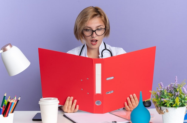 Zaskoczony młoda lekarka w szacie medycznej ze stetoskopem i okularami siedzi przy stole z narzędziami medycznymi, trzymając i patrząc na folder na białym tle na niebieskim tle