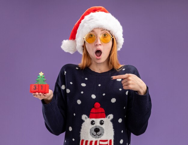 Zaskoczony, młoda piękna dziewczyna ubrana w świąteczny sweter i kapelusz w okularach trzyma i wskazuje na świąteczne zabawki na białym tle na fioletowym tle