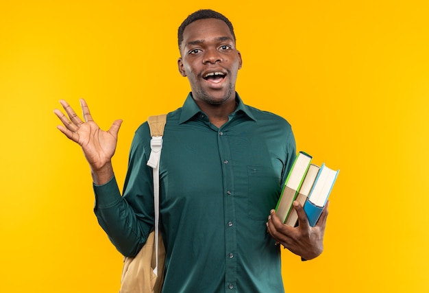 Zaskoczony młody afroamerykański student z plecakiem trzymającym książki i trzymającym rękę otwartą na białym tle na pomarańczowej ścianie z miejscem na kopię
