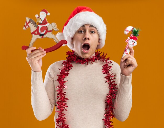 Zaskoczony młody facet w świątecznym kapeluszu z girlandą na szyi, trzymający świąteczną zabawkę z cukierkiem na żółtym tle