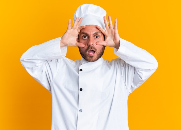 Zaskoczony młody kaukaski kucharz w mundurze szefa kuchni i czapce, patrzący na kamerę, robiący duże oczy izolowane na pomarańczowej ścianie
