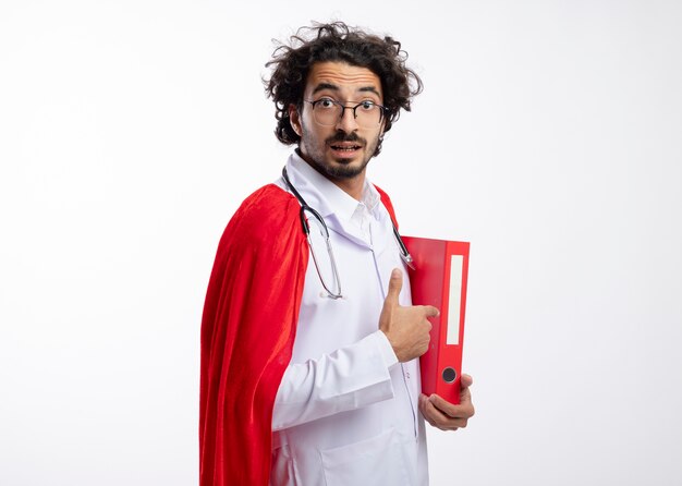 Zaskoczony młody kaukaski mężczyzna w okularach optycznych w mundurze lekarza z czerwonym płaszczem i stetoskopem na szyi stoi bokiem, trzymając i wskazując na folder plików z miejscem na kopię
