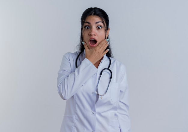 Zaskoczony, młody lekarz kobiet na sobie szlafrok medyczny i stetoskop, patrząc, kładąc rękę na brodzie na białym tle