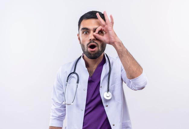 Zaskoczony, młody lekarz płci męskiej sobie stetoskop medycznej sukni pokazujący wygląd gest na na białym tle biały