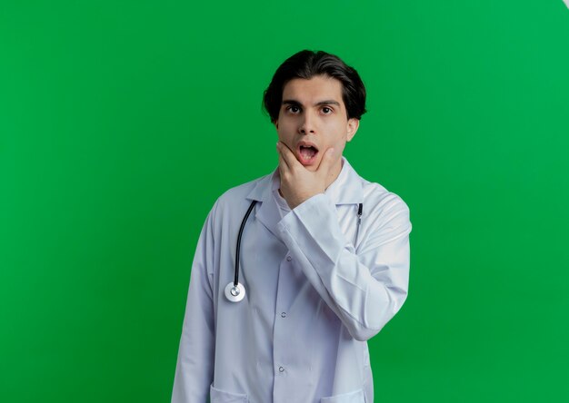 Zaskoczony młody lekarz płci męskiej ubrany w szlafrok i stetoskop trzymając podbródek
