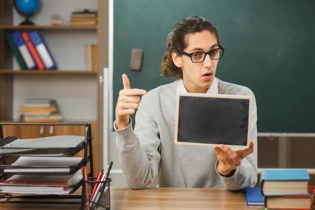 zaskoczony młody nauczyciel płci męskiej w okularach, siedzący przy biurku, trzymający i wskazujący na mini tablicę z narzędziami szkolnymi włączonymi w klasie