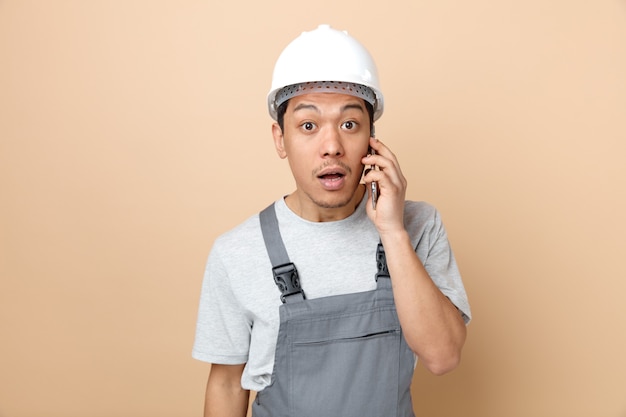 Zaskoczony, młody pracownik budowlany w kasku i mundurze rozmawia przez telefon