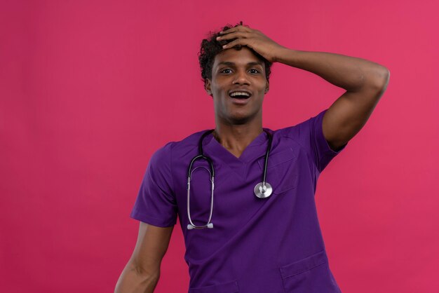 Zaskoczony, młody, przystojny, ciemnoskóry lekarz z kręconymi włosami w fioletowym mundurze ze stetoskopem trzymający rękę na głowie