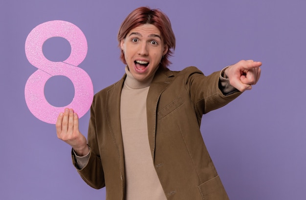 Bezpłatne zdjęcie zaskoczony młody przystojny mężczyzna trzymający różową cyfrę osiem i wskazujący na bok
