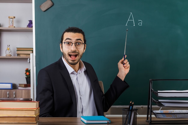 Zaskoczony nauczyciel w okularach wskazuje na tablicę kijem wskaźnikowym siedzącym przy stole z narzędziami szkolnymi w klasie