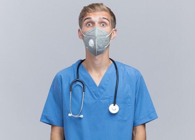 Zaskoczony patrząc na kamery młody mężczyzna lekarz ubrany w mundur lekarza ze stetoskopem i maską medyczną na białym tle na białej ścianie
