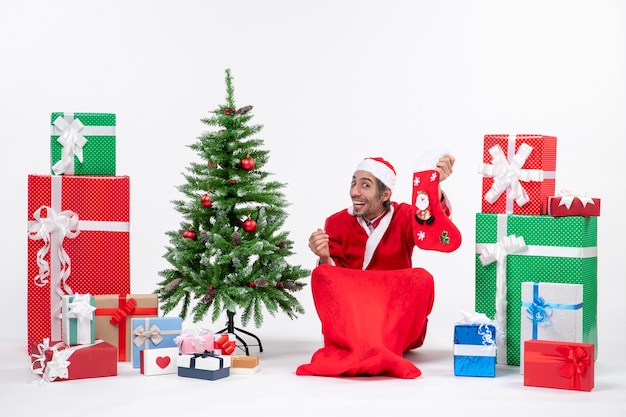 Zaskoczony Pozytywny Podekscytowany święty Mikołaj Siedzi Na Ziemi I Trzyma świąteczną Skarpetę W Pobliżu Prezentów I Udekorowane Drzewo Noworoczne Na Białym Tle