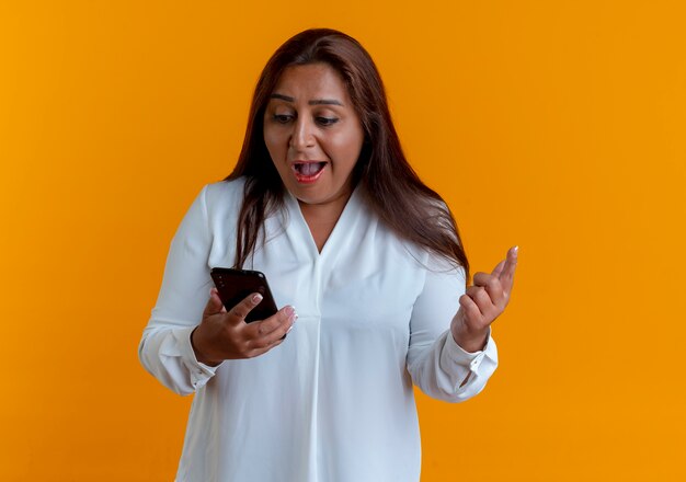 Zaskoczony przypadkowy kaukaski kobieta w średnim wieku trzymając i patrząc na telefon na białym tle na żółtej ścianie