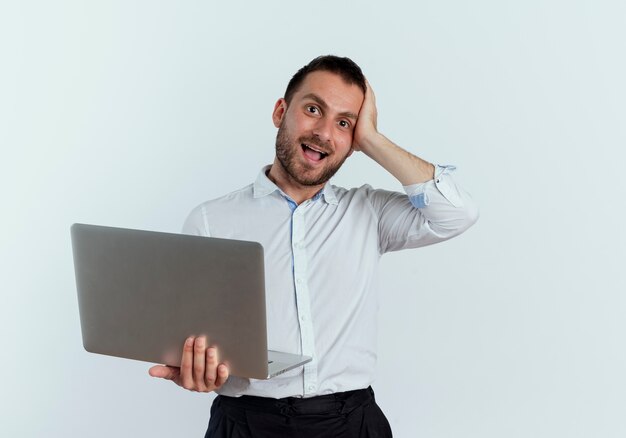 Zaskoczony, przystojny mężczyzna kładzie rękę na głowie trzymając laptopa na białym tle na białej ścianie