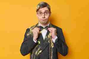 Bezpłatne zdjęcie zaskoczony zabawny mężczyzna dopasowuje muszkę ubraną w formalny strój nosi okrągłe sukienki okularowe na specjalną okazję posmarowane serpantynowym sprayem izolowanym na żółtym tle przygotowania do wakacji