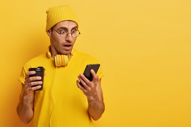 zaskoczony zszokowany mężczyzna wpatruje się w smartfon, nosi przezroczyste okulary i żółty kapelusz, pije kawę na wynos, zdumiony okropnymi wiadomościami w internecie. Monochromatyczny, kolor żółty