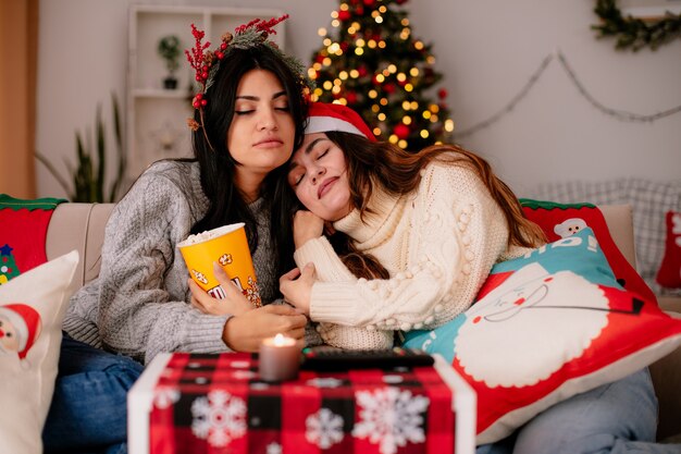 zaspane ładne młode dziewczyny w czapce Mikołaja i wieńcu ostrokrzewu trzymają wiaderko popcornu siedząc na fotelach i ciesząc się świętami w domu