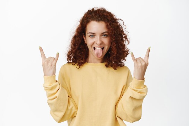 Zaszalejmy. Odważna podekscytowana rudowłosa dziewczyna pokazująca gest języka i heavy metalowych rogów, ciesząca się imprezą, bawiąca się, stojąca na białym tle