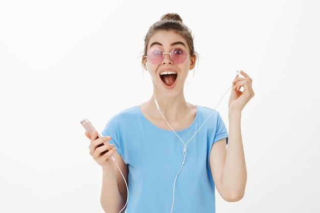 Zbliżenie: atrakcyjna nowoczesna kobieta w okularach przeciwsłonecznych, słuchanie muzyki, słuchanie piosenki w słuchawkach, trzymając smartfon